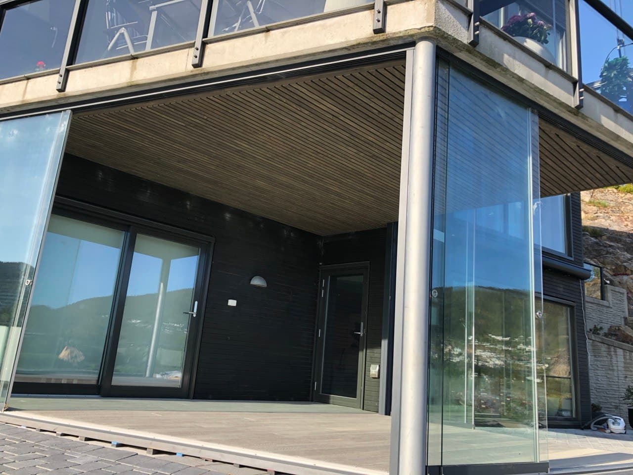 Innglasset uteplass under balkong med mørk kledning og spiletak i tre